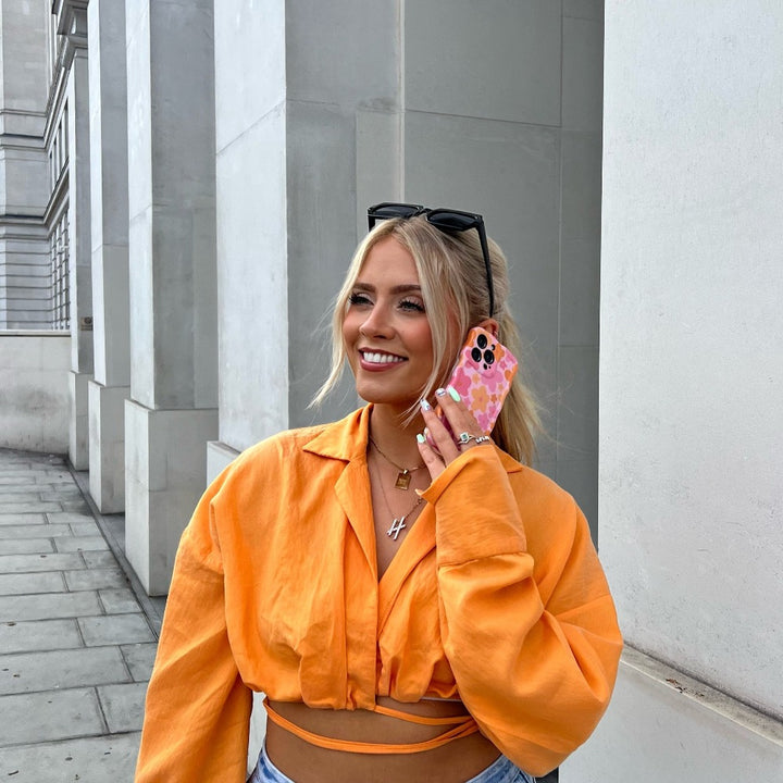 A woman with long blonde hair, wearing a bright orange cropped shirt and light blue jeans, stands on a sidewalk smiling while holding a phone with an extra protective Wavy Phone Case - Pink & Orange Bloom by Coconut Lane to her ear. She has sunglasses perched on her head and a necklace with a capital letter "H".