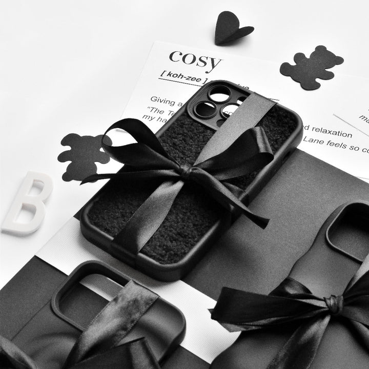 A monochrome image featuring the Cosy Teddy Phone Case - Black by Coconut Lane, tied with a black ribbon and placed on a black-and-white textured surface. Also shown are a dictionary definition of "cosy," black teddy bear cutouts, and the letter "B.