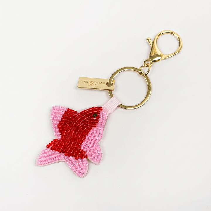A beaded fish-shaped keyring in shades of pink and red, attached to a gold metal split ring a clasp, shown on a plain white background.