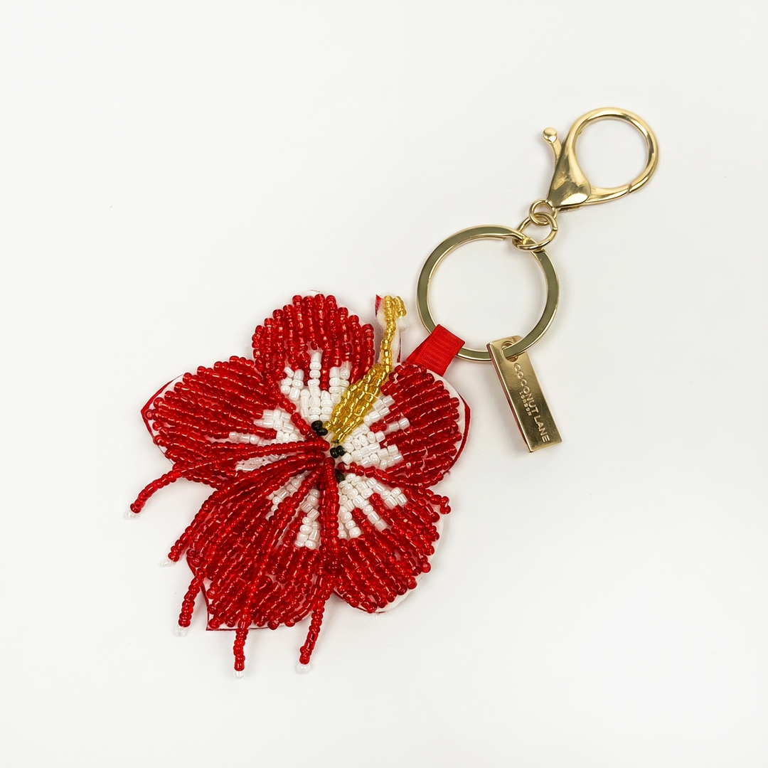 A beaded keyring featuring a red and white hibiscus flower with gold accents, attached to a gold split ring and clasp on a white background.