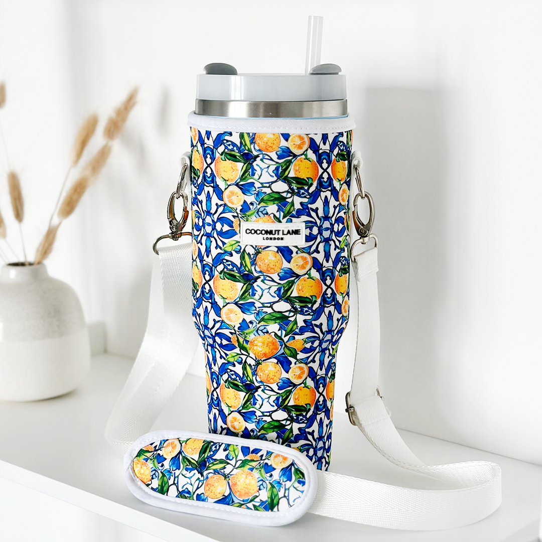 A vibrant Coconut Lane Tumbler Carry Case - Amalfi Lemon, adorned with a charming floral and fruit pattern featuring lemons and green leaves on a blue background. It includes a coordinated strap and an attached small pouch. In the background, there is a decorative vase filled with dried wheat stalks resting on a white surface.