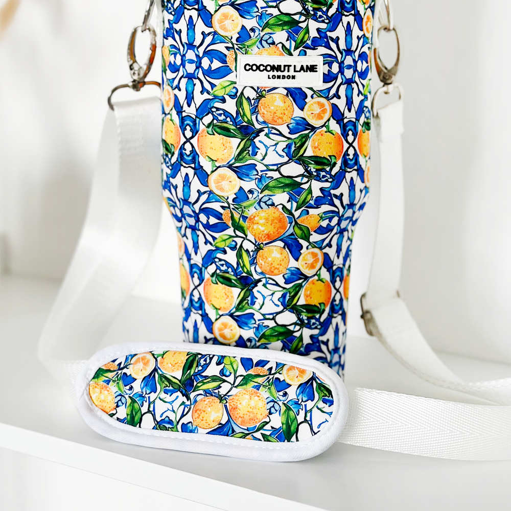The "Tumbler Carry Case - Amalfi Lemon" by Coconut Lane is a stylish sling bag featuring a vibrant blue and white floral and lemon print, perfect for carrying your Stanley Quencher on the go. It includes white adjustable straps with silver clasps and a padded shoulder strap adorned with the same eye-catching pattern.