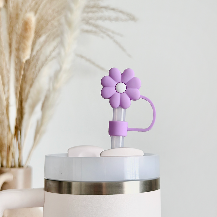 A close-up of a white tumbler with a silver rim, showcasing the Coconut Lane Lilac Flower Straw Cover. In the background, ornamental pampas grass stems are placed in a vase, slightly blurred to enhance the light and airy ambiance.