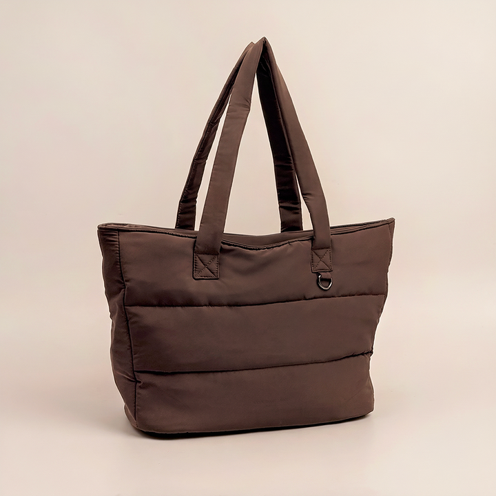 Chocolate brown padded tote bag with quilted panels, long shoulder straps and metal d-ring detail, photographed on a nude beige background.