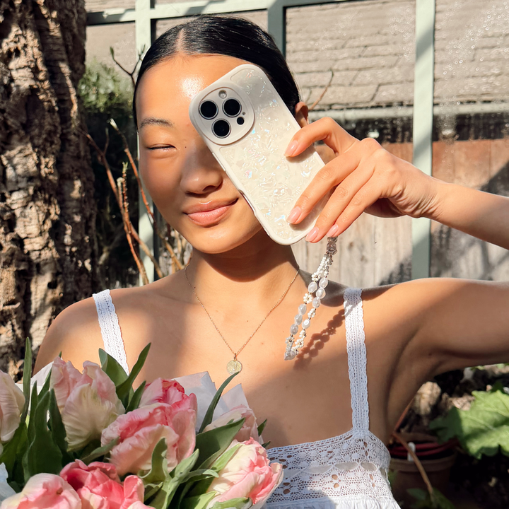Girl in a greenhouse holding flowers posing with her Moonstone Pearl Phone Case that's shining in the sun