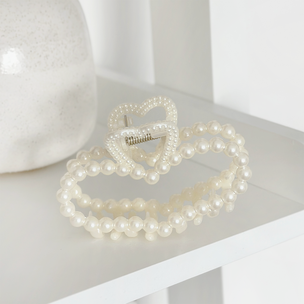 A Pearl Heart Claw Clip from Coconut Lane, beautifully decorated with pearl-like beads, rests on a white shelf. This minimalist and elegant hair accessory features two intertwined heart shapes embellished with small, white pearls.