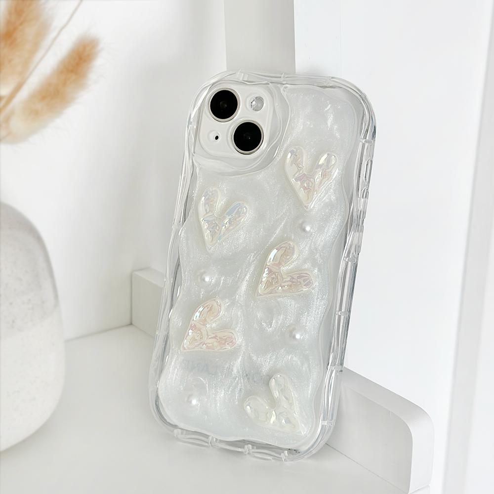 The Pearly Hearts Phone Case - Clear by Coconut Lane is an iridescent phone case with a textured design featuring raised, shimmering hearts and small pearl-like embellishments. This extra protective case is propped up on a white surface next to a vase containing dried wheat stalks.