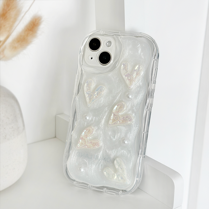 The Pearly Hearts Phone Case - Clear by Coconut Lane is an iridescent phone case with a textured design featuring raised, shimmering hearts and small pearl-like embellishments. This extra protective case is propped up on a white surface next to a vase containing dried wheat stalks.