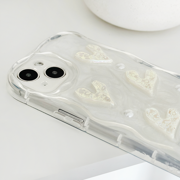 A smartphone with a dual-camera setup is placed on a white surface. The phone is encased in Coconut Lane's Pearly Hearts Phone Case - Clear, featuring a pearly, wavy texture and raised designs of hearts. The image focuses on the back of the phone, highlighting the camera and the details of the Pearly Hearts Phone Case - Clear.