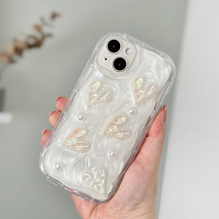 A hand holds an iPhone15 with an extra Coconut Lane Pearly Hearts Phone Case - Clear. The case showcases a pearlescent white design with raised heart shapes and small pearl-like embellishments. The phone is held in front of a blurred background.
