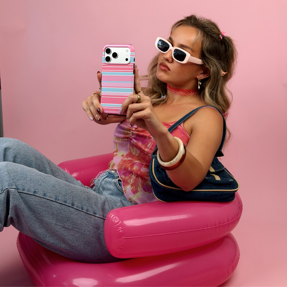 A woman wearing pink sunglasses, a pink floral top, and blue jeans sits on a pink inflatable chair, holding a striped pink, blue and white phone case against a solid pink background.