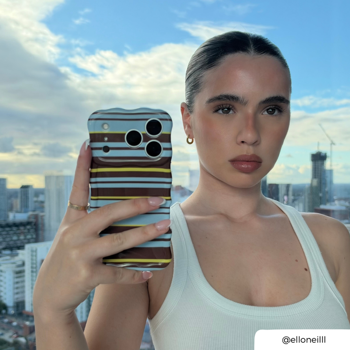 Woman taking a mirror selfie, holding an iphone in a brown, blue and yellow horizontal striped wavy phone case.