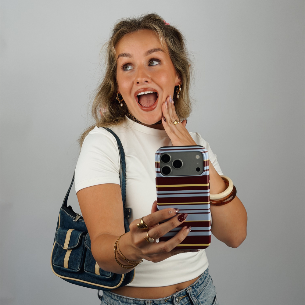 A woman in a white crop top and denim shorts holds up a brown, blue and yellow striped phone case, looking surprised and smiling. She is wearing a blue denim shoulder bag and gold jewellery against a plain white studio background.