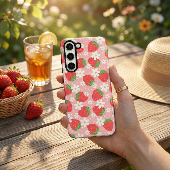 Hand holding Coconut Lane Samsung phone case with pink strawberry design; iced tea, strawberries, and hat in background.