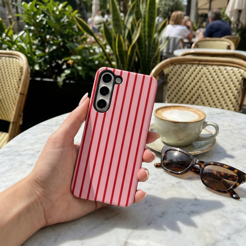 Hand holding a Samsung Tough Phone Case with red and white cherry stripe design, near coffee cup and sunglasses on outdoor table.