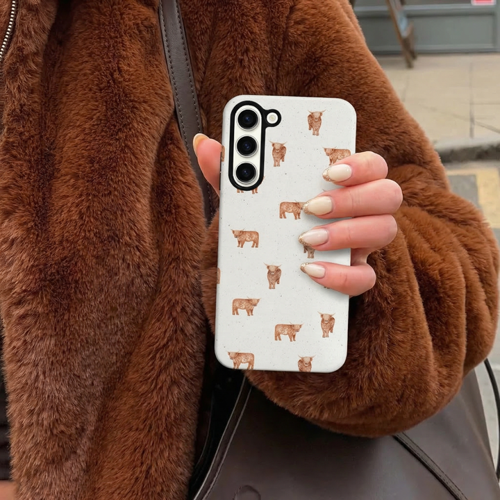 Person in a brown faux fur coat holding a Samsung phone case with a Highland Cow illustration by Coconut Lane.