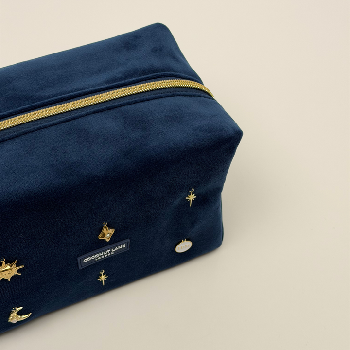 Stardust After Dark makeup bag. Dark navy blue velvet material with gold zip, and sewn-on gold celestial-themed charms including a sun, moon, star, diamond shape and 11:11 charm.  