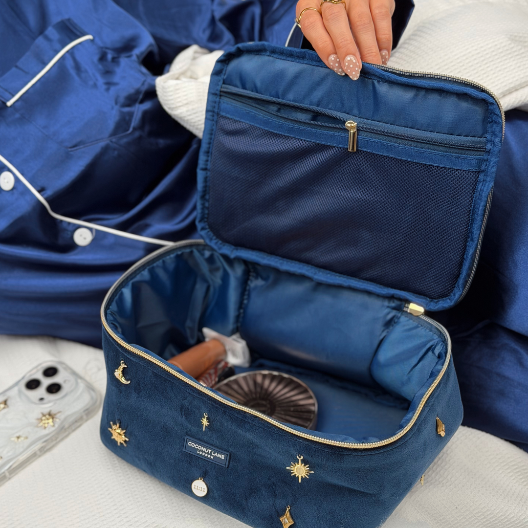 Woman wearing satin navy blue pyjamas. She is opening a stardust After Dark Vanity Case. A dark blue navy velvet vanity case with gold zip, and sewn-on gold celestial-themed charms. Including a moon, sun, star, diamond shape and 11:11 charm. 
