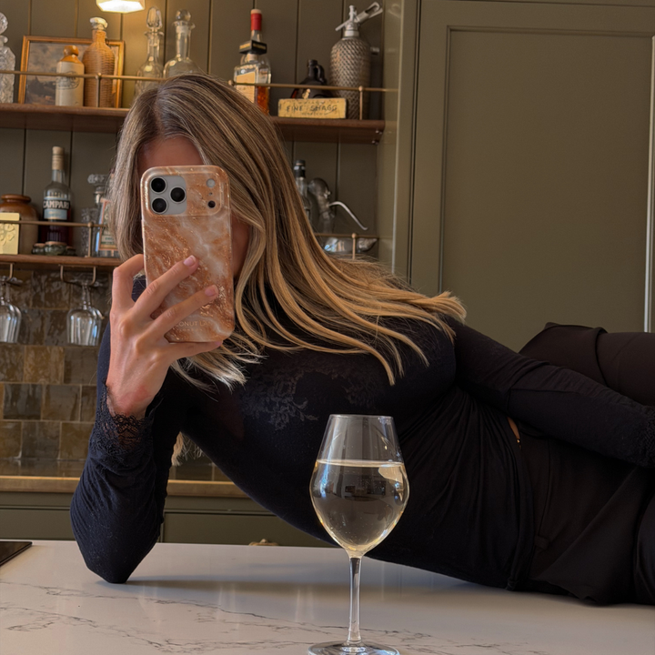 Woman with blonde hair wearing a black long sleeve top is holding an iphone in a marble effect phone case with cream, gold, light brown and white tones running through it.