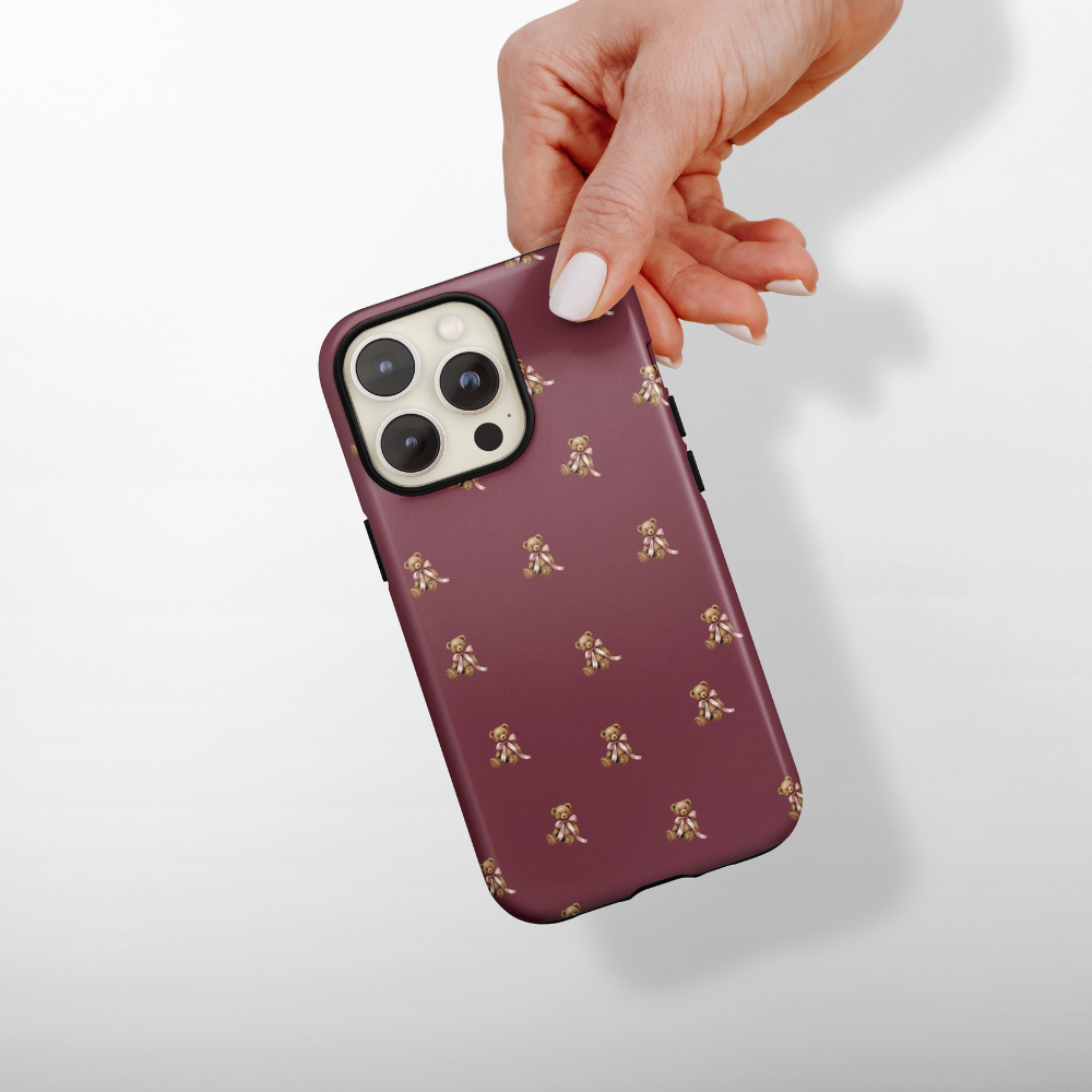 Hand holding a phone with a maroon case featuring a teddy pattern on a light gray background