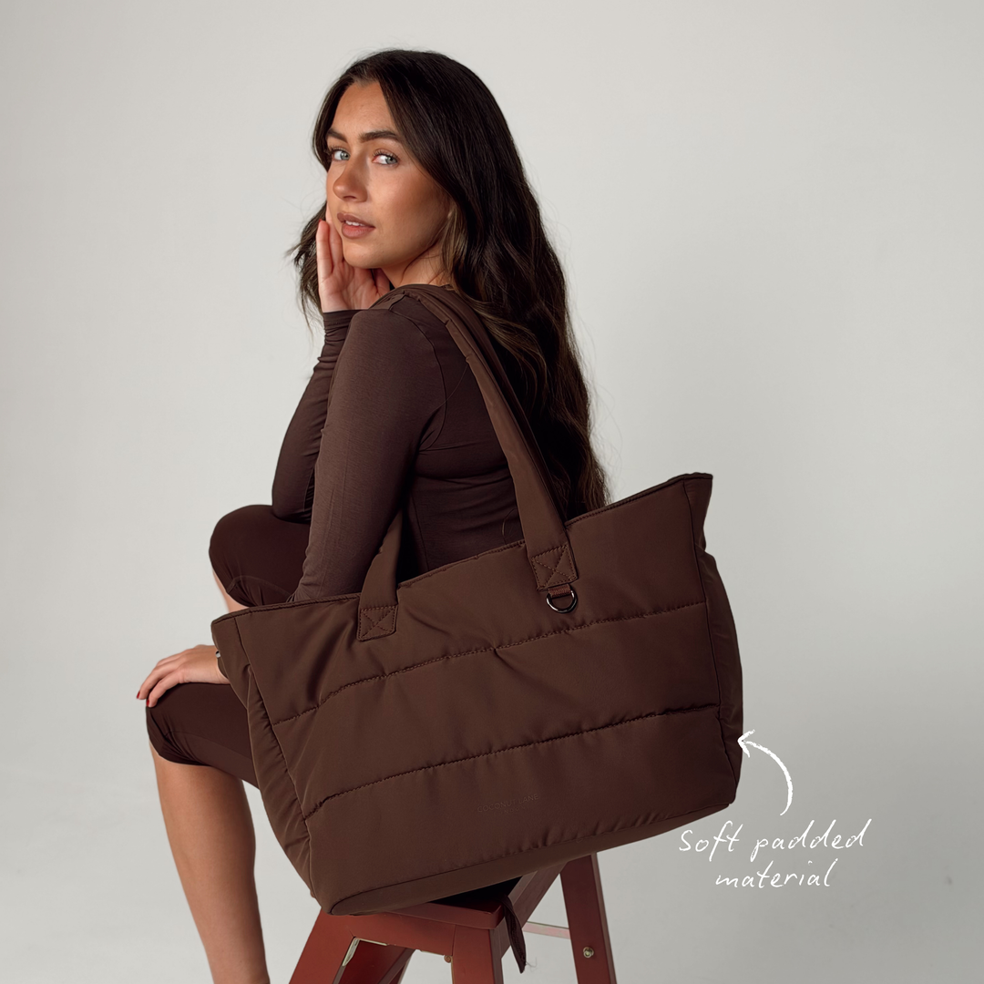 Woman with brunette hair, wearing a brown pilates outfit. She has the Main Carry Choc Brown Puffer Bag on her shoulder. The image annotates the soft padded material of the bag. 