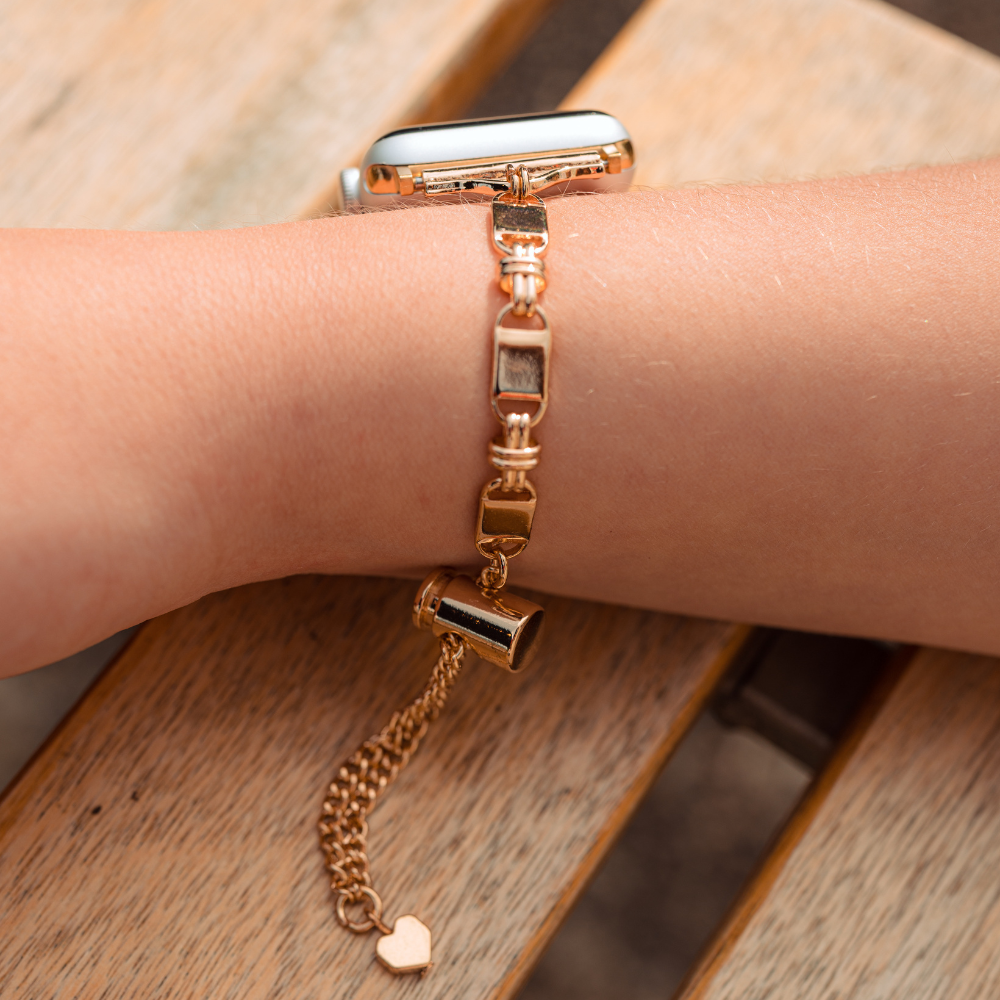 Close-up of a person's wrist wearing the Simple Gold Bracelet Apple Watch Strap by Coconut Lane. The bracelet showcases rectangular segments separated by intricate links, reminiscent of an elegant Apple Watch strap, and features a dangling chain ending in a small heart charm. The background appears to be a wooden surface.