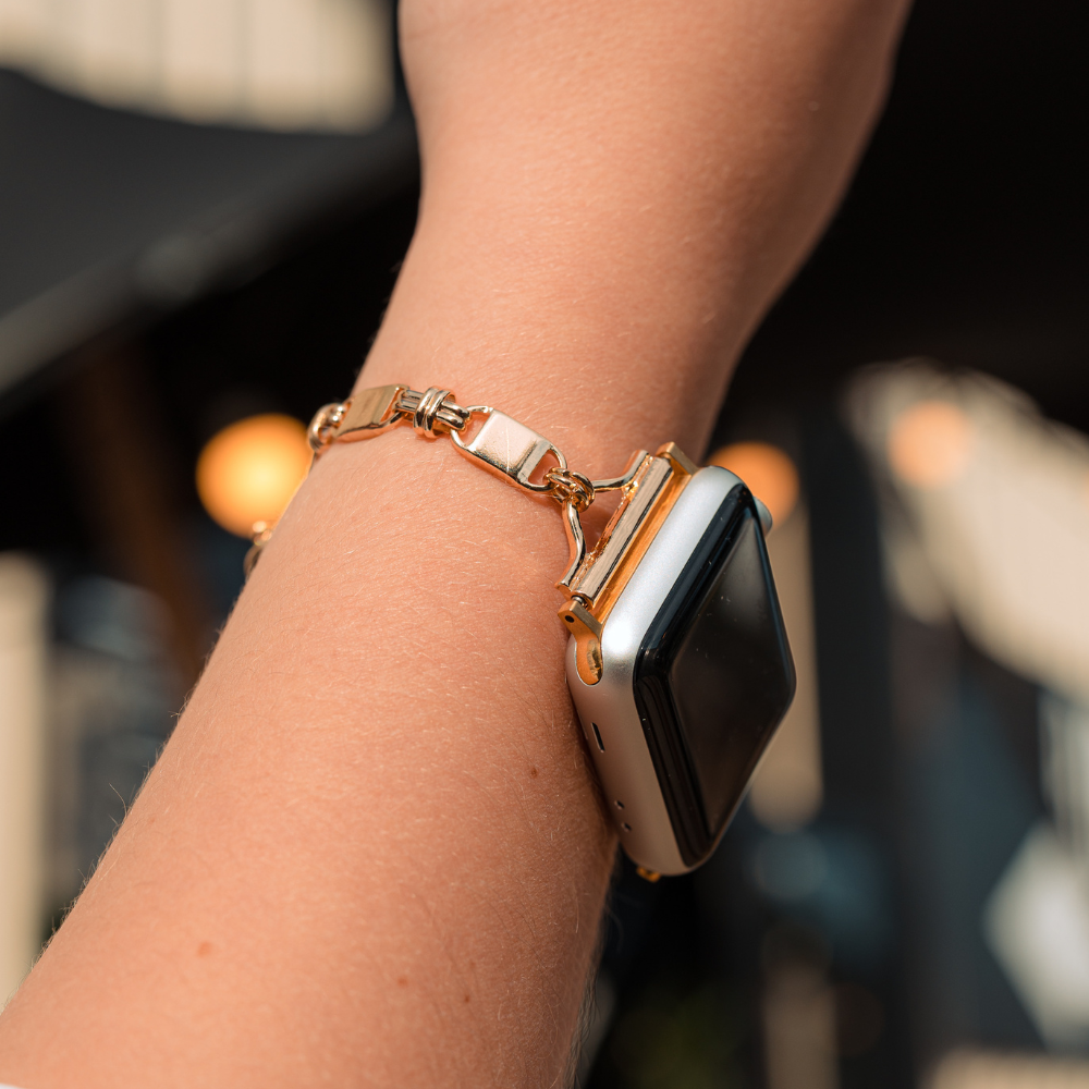 A close-up of a person's wrist wearing the Simple Gold Bracelet Apple Watch Strap by Coconut Lane. The person has a light skin tone, and the background is blurred with out-of-focus lights and architectural details.