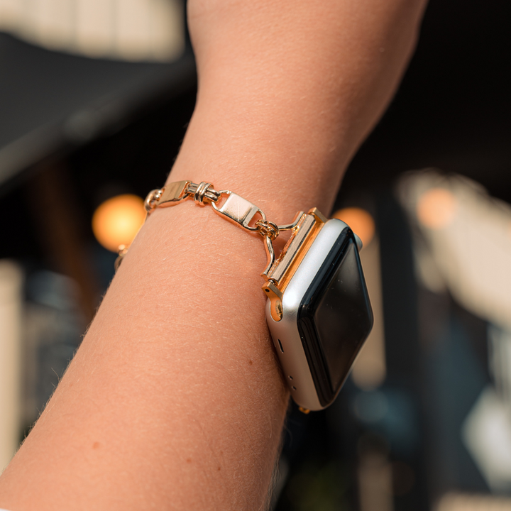 A close-up of a person's wrist wearing the Simple Gold Bracelet Apple Watch Strap by Coconut Lane. The person has a light skin tone, and the background is blurred with out-of-focus lights and architectural details.
