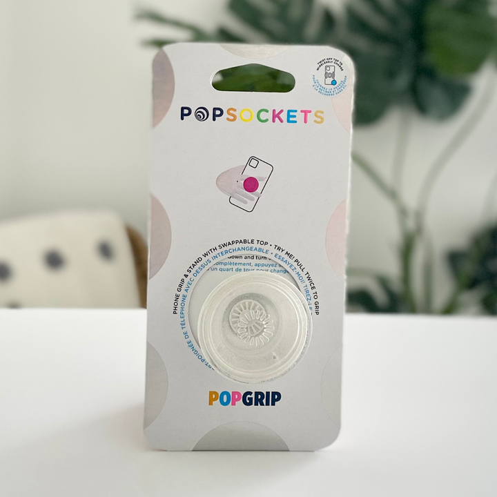 A packaged Coconut Lane PopSocket Grip - Clear Silver Glitter is displayed on a white surface with a blurred plant in the background. The packaging shows the product inside, featuring a white circular grip with a simple floral design and clear silver glitter accents. The Coconut Lane logo is visible at the top.