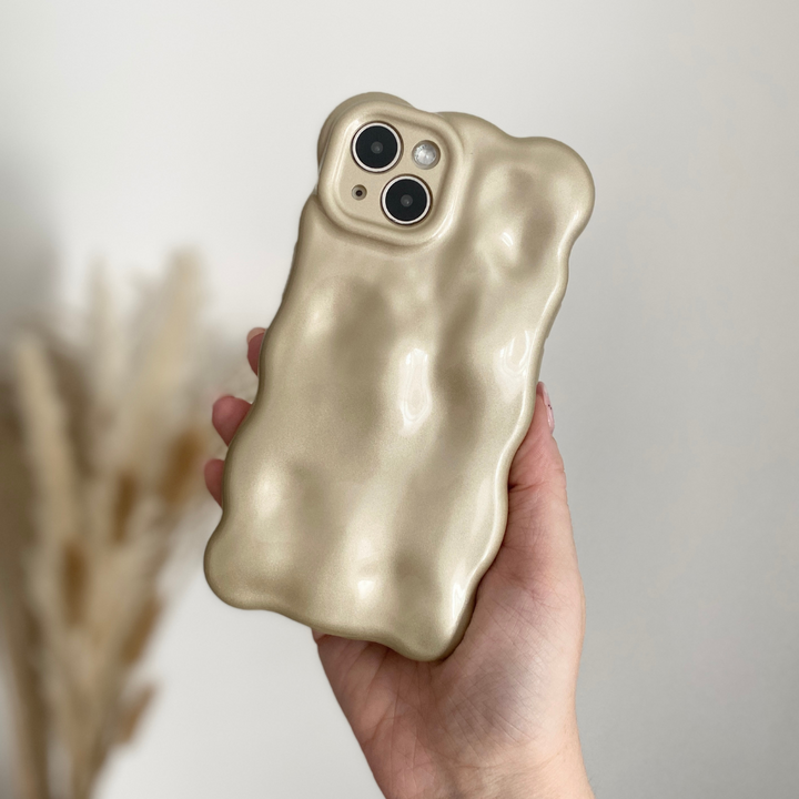 A person is holding a beige-colored iPhone 15 with Coconut Lane's distinctive 3D Pearl Phone Case - Gold. The softly blurred background features dried pampas grass, highlighting the soft and cushiony texture of the gold-accented case.