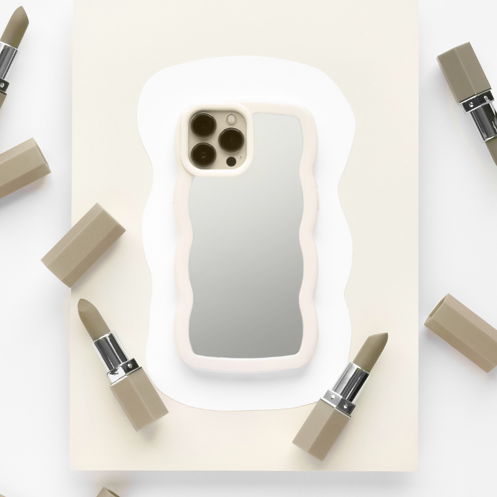 A sleek and stylish iPhone adorned with the Coconut Lane Curvy Mirror Phone Case in Cream is centered on a piece of off-white paper. The phone's camera is visible, reflecting off the back of the case. Surrounding the device are six muted beige lipsticks, some with their caps removed, arranged in a circular pattern.
