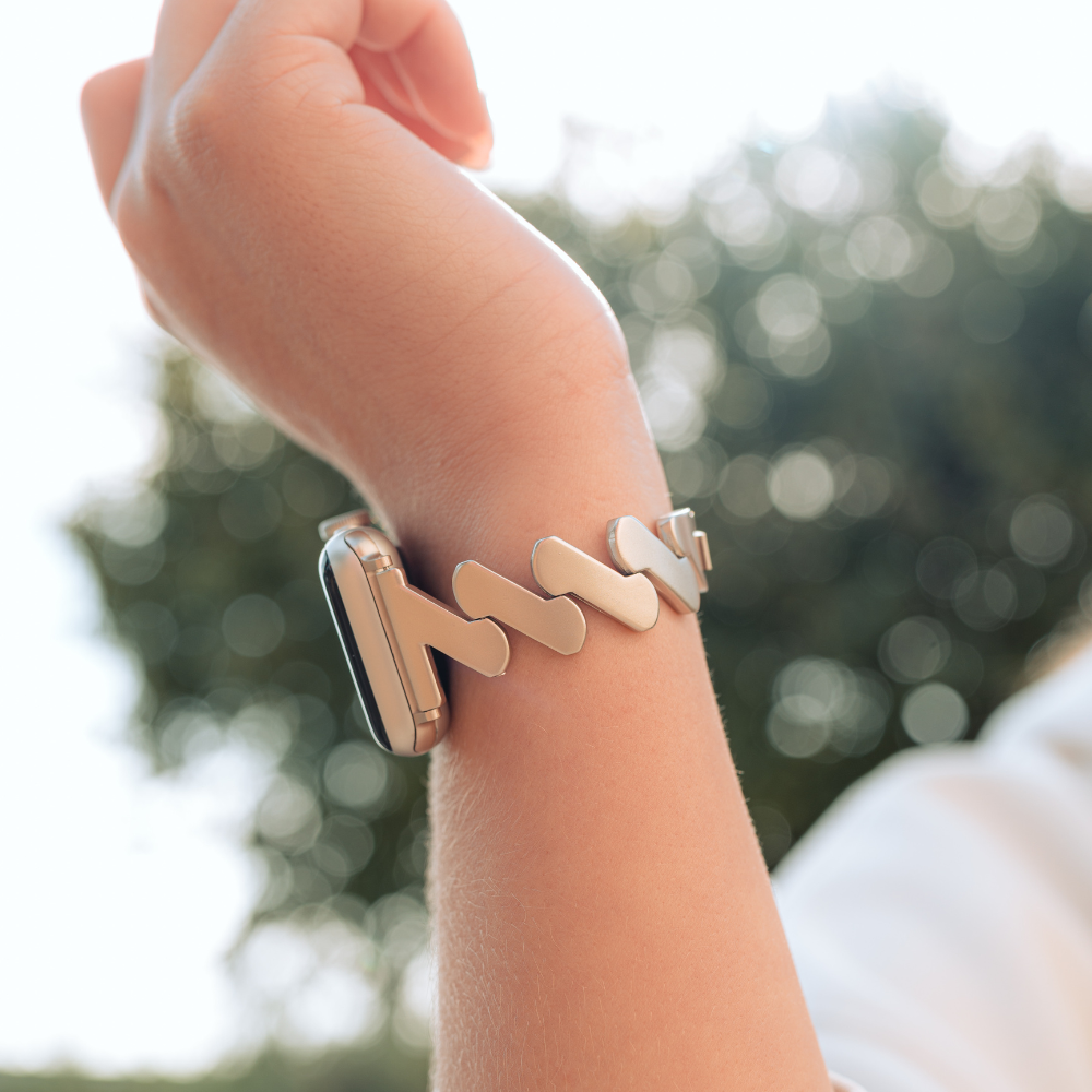 Close-up of a person's wrist wearing Coconut Lane's stylish, modern Get Wavy Apple Watch Strap - Starlight. The background is slightly blurred, showing an outdoor setting with trees and natural light. The Stainless Steel Metal Adjustable Apple Watch Strap fits ALL Apple Watch models, showcasing the look prominently.