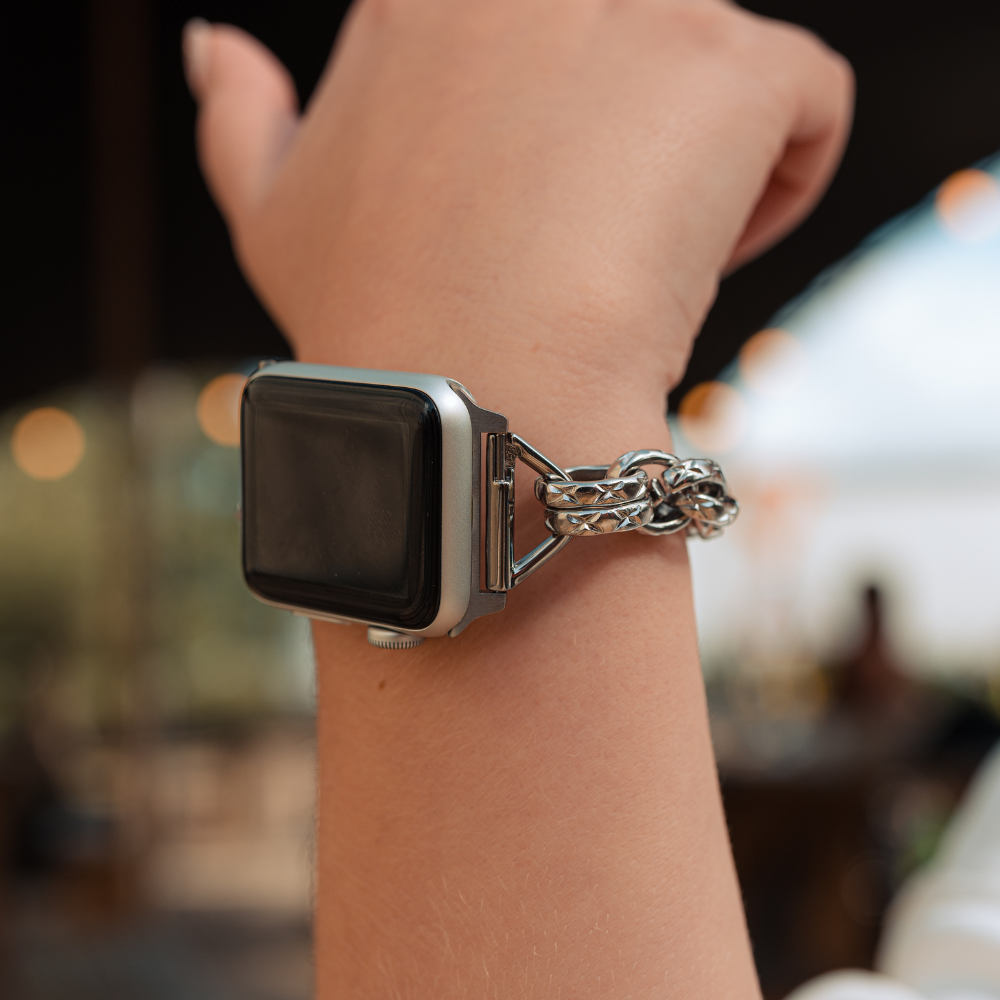 A close-up of a person's wrist wearing the Chain Bracelet Apple Watch Strap - Silver by Coconut Lane, paired with a unique silver chain bracelet. The background shows a softly blurred indoor setting, suggesting a casual environment.