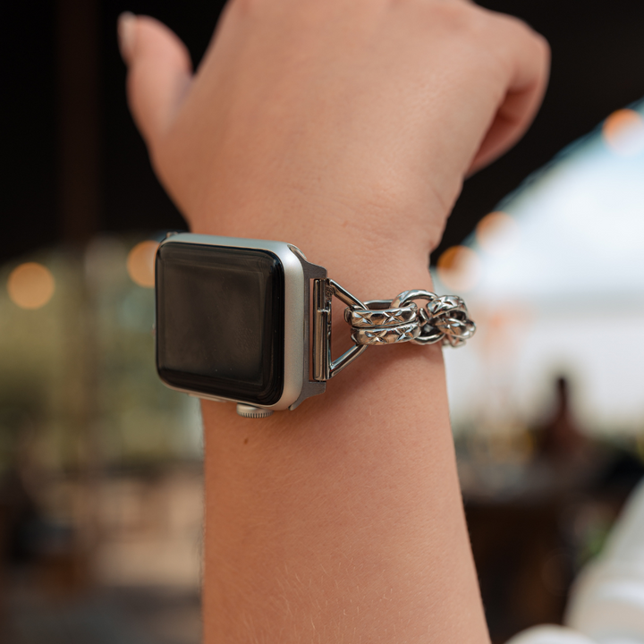 A close-up of a person's wrist wearing the Chain Bracelet Apple Watch Strap - Silver by Coconut Lane, paired with a unique silver chain bracelet. The background shows a softly blurred indoor setting, suggesting a casual environment.