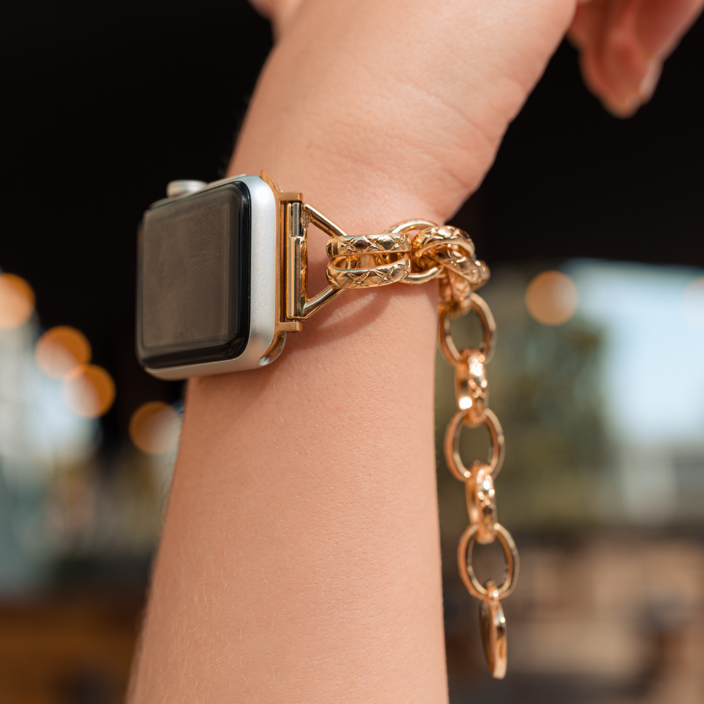 Close-up of a wrist wearing the Coconut Lane Chain Bracelet Apple Watch Strap - Gold, featuring a metallic gold chain strap. The background is blurred, with ambient lighting creating bokeh effects. The hand is slightly raised, prominently showcasing the watch strap.