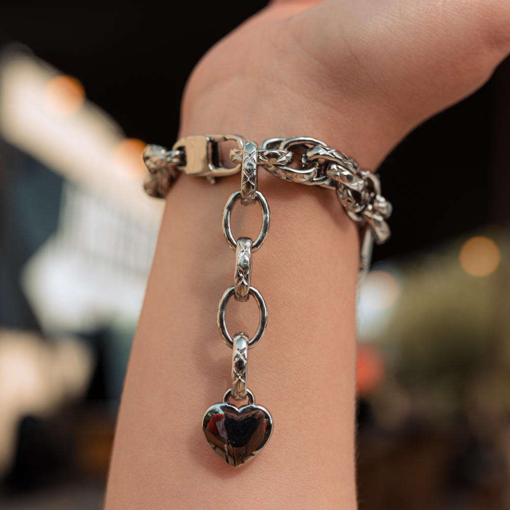 A Chain Bracelet Apple Watch Strap - Silver by Coconut Lane, showcasing its intricate links and attached heart-shaped charm, is elegantly worn on a person's wrist. The blurred background with soft lights and indistinct objects accentuates the bracelet's shiny stainless steel design.