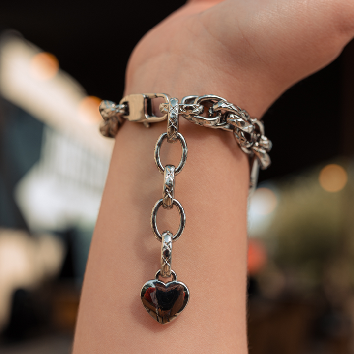 A Chain Bracelet Apple Watch Strap - Silver by Coconut Lane, showcasing its intricate links and attached heart-shaped charm, is elegantly worn on a person's wrist. The blurred background with soft lights and indistinct objects accentuates the bracelet's shiny stainless steel design.