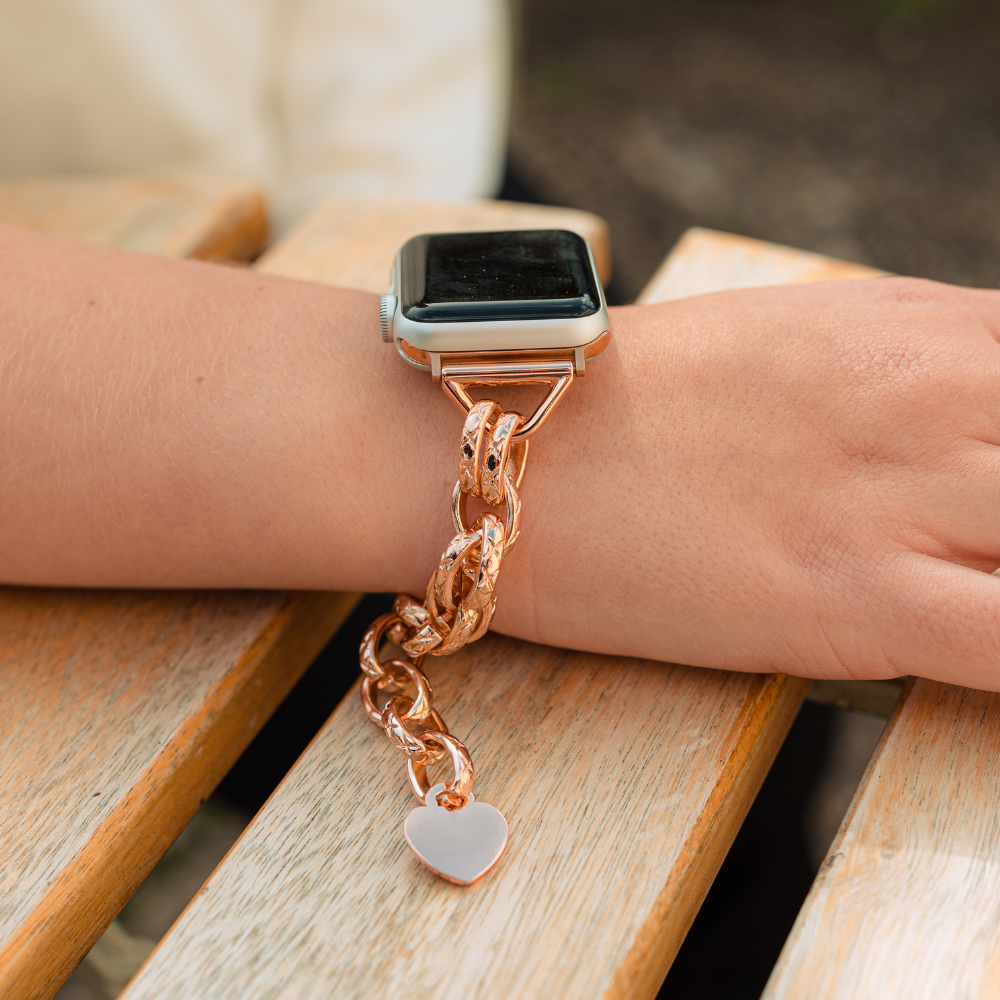 A person is wearing Coconut Lane's Chain Bracelet Apple Watch Strap in Rose Gold, which includes a heart-shaped charm. The arm rests on a wooden surface against a blurred background. This stainless steel strap features interlocking links with sparkling embellishments and is adjustable for the perfect fit.