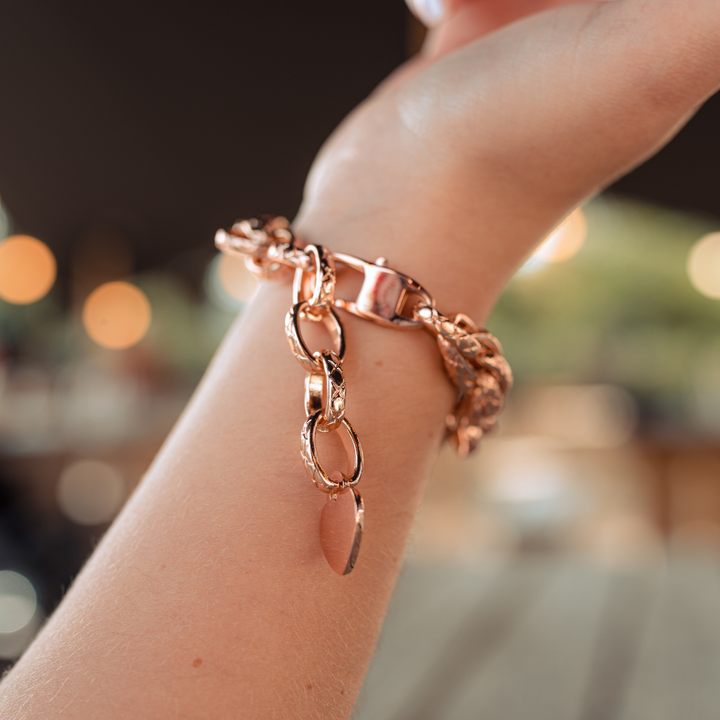 A close-up of a person's wrist wearing the Chain Bracelet Apple Watch Strap - Rose Gold by Coconut Lane. The strap features large interlocking links and a dangling charm. The background is softly blurred with warm bokeh lighting, suggesting an indoor setting.