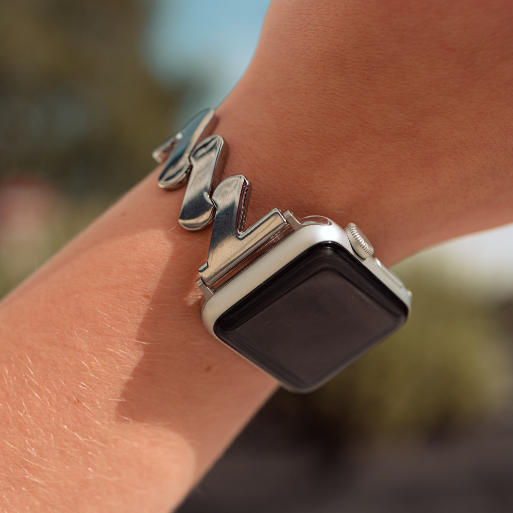 A close-up shot features a wrist adorned with the Coconut Lane Get Wavy Apple Watch Strap in Silver. The Apple Watch's screen is black and turned off, set against a blurred outdoor backdrop highlighting vibrant greenery.