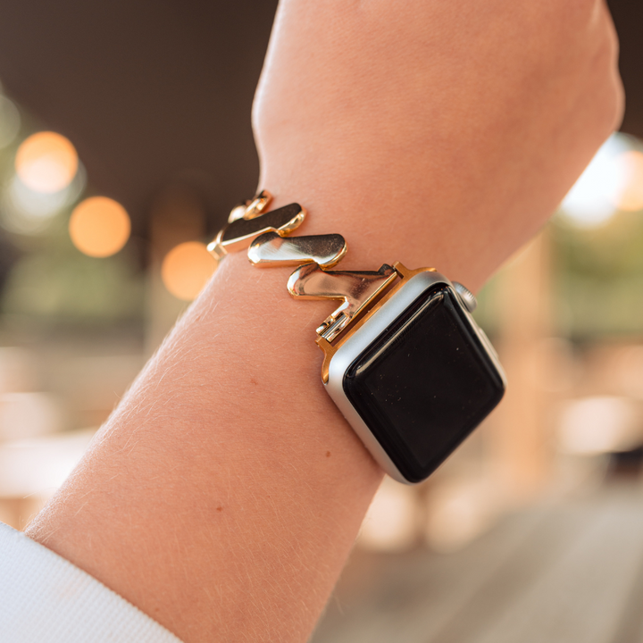 A wrist adorned with an Apple Watch featuring the Get Wavy Apple Watch Strap - Gold by Coconut Lane. The sleek, adjustable band boasts a metallic wave pattern. The blurred background, illuminated with warm lights, provides a cozy ambiance.
