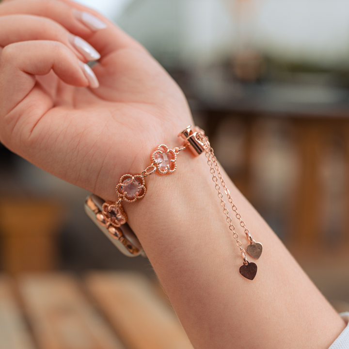 A close-up of a person's wrist adorned with Coconut Lane's Rose Gold Flower Bracelet Apple Watch Strap, showcasing its floral-shaped links and small dangling heart charms. The person's relaxed hand flaunts manicured nails, with a blurred outdoor background enhancing the elegance of the look.