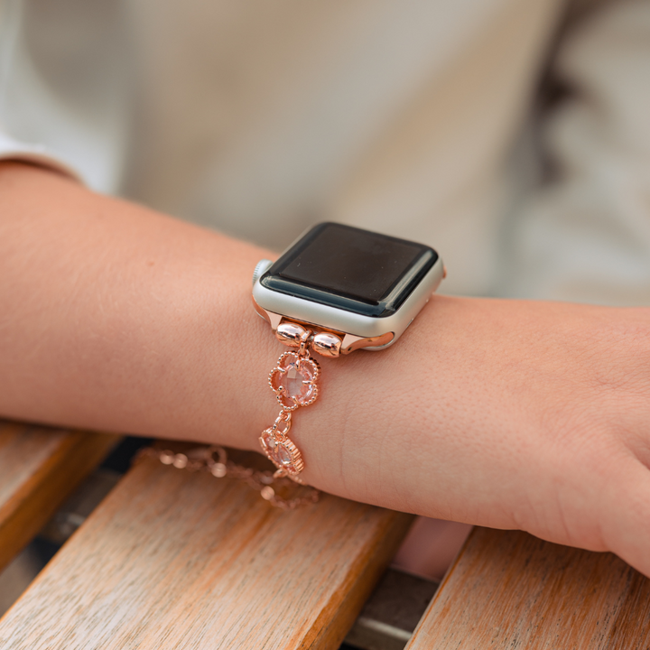 A close-up of a person's wrist wearing a smartwatch with the Coconut Lane Rose Gold Flower Bracelet Apple Watch Strap. The stainless steel chain strap glistens as the arm rests on a wooden surface, and they are dressed in a light-colored sleeve.