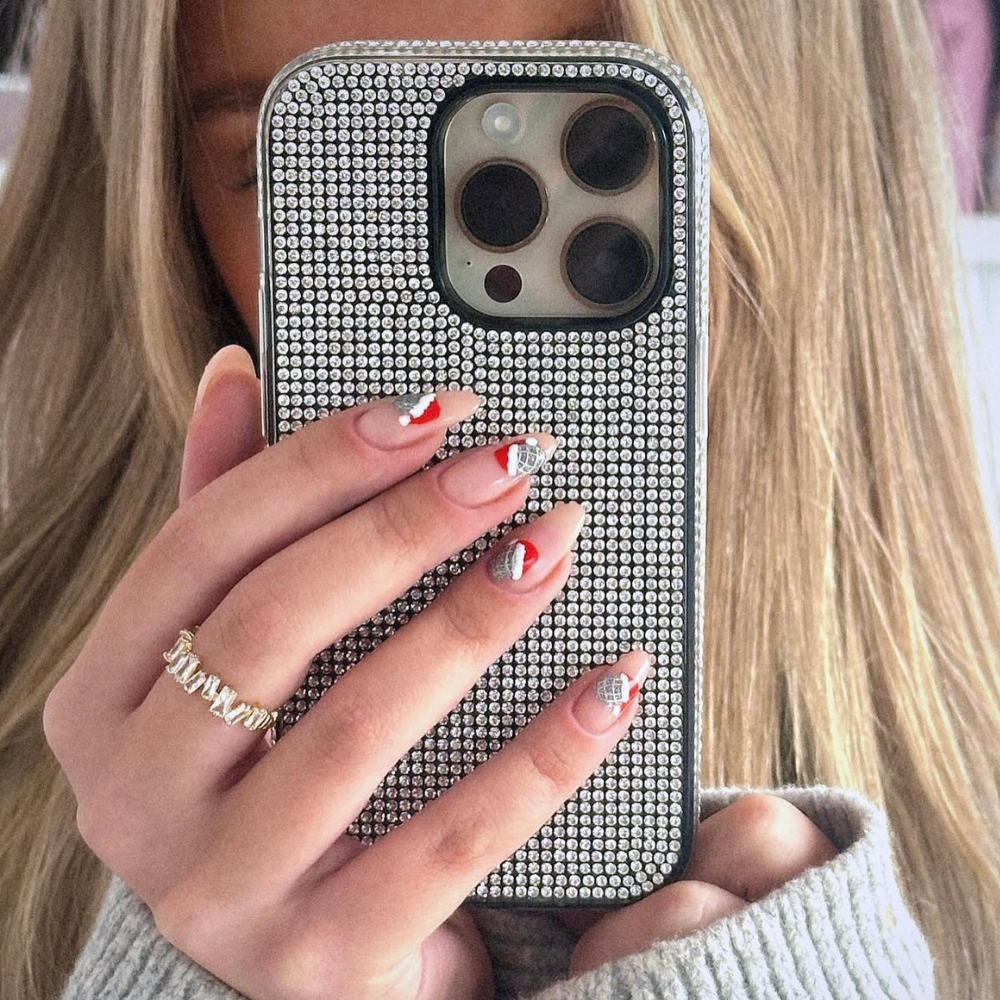 A person with long, blonde hair is holding a smartphone with the Coconut Lane Disco Sparkle Phone Case - Silver up to their face. Their nails are manicured with various designs, including red and white patterns, and they are wearing a gold ring adorned with multiple small stones.
