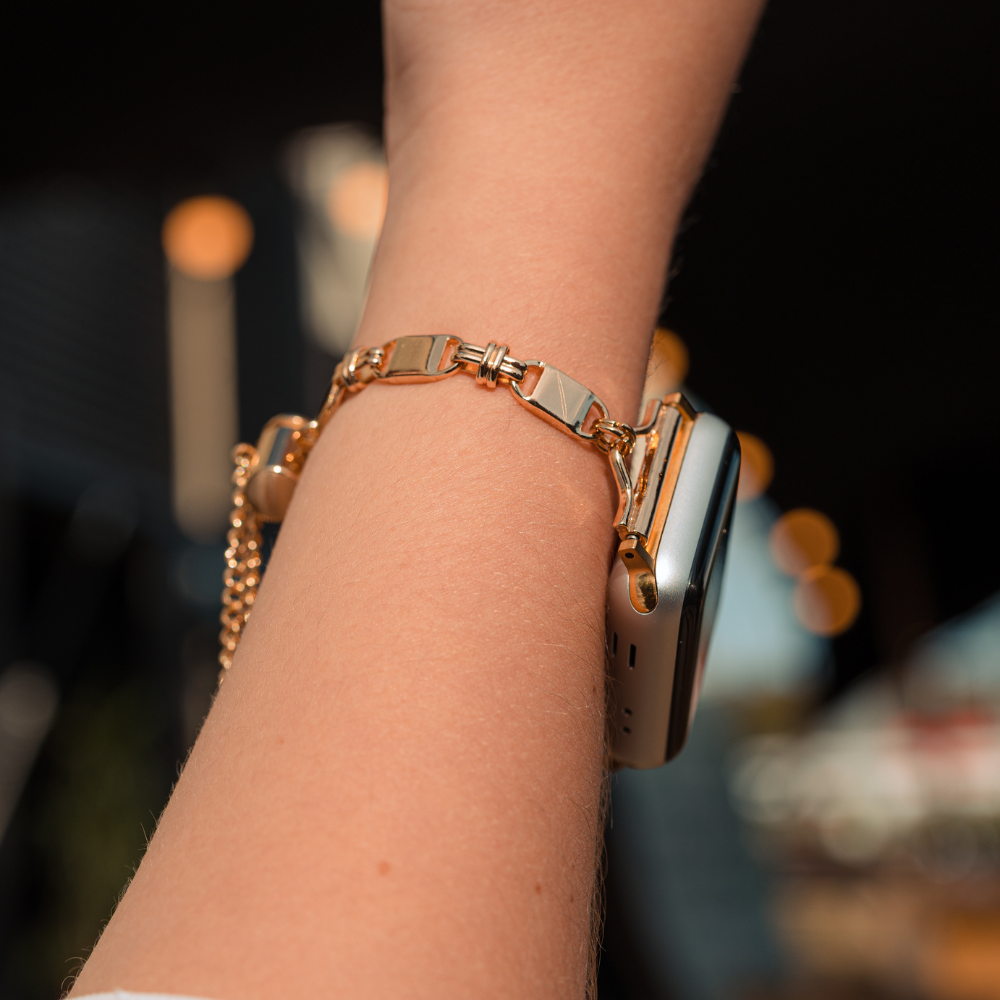 A close-up of a person's wrist adorned with a Simple Gold Bracelet Apple Watch Strap from Coconut Lane. The background is blurred, featuring warm-toned bokeh lights and some indistinct shapes.