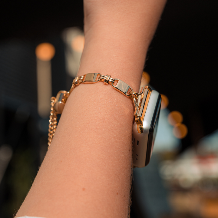 A close-up of a person's wrist adorned with a Simple Gold Bracelet Apple Watch Strap from Coconut Lane. The background is blurred, featuring warm-toned bokeh lights and some indistinct shapes.