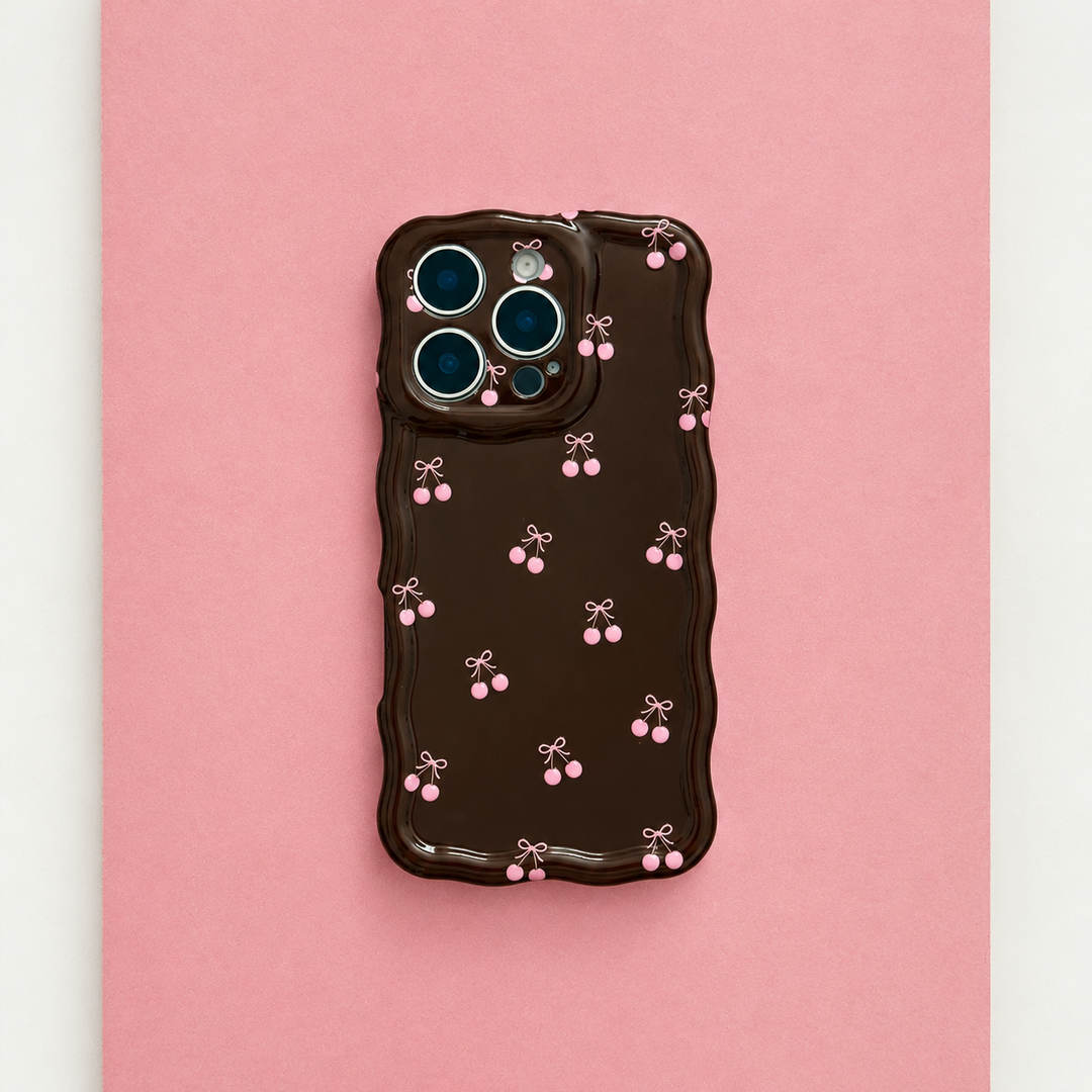 Wavy Phone Case featuring a glossy chocolate brown finish with delicate pink cherry and bow illustrations, wavy edges and triple camera cutouts, photographed on a pink background.