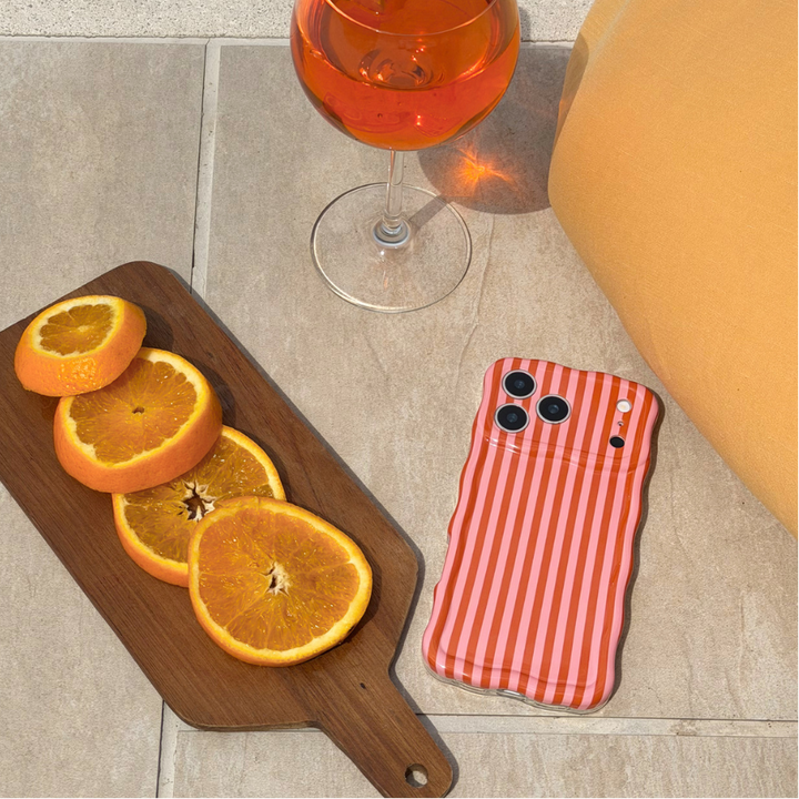 Phone case with orange and pink striped pattern next to sliced oranges and a glass of orange juice on a stone surface.