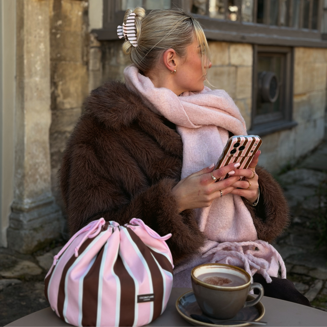 Woman wearing a baby pink scarf and brown faux fur jacket, holding a wavy cocoa and pink stripe phone case. On the table beside her is a drawstring toiletry bag featuring chocolate brown, baby pink and white stripes with pink drawstring closure. 