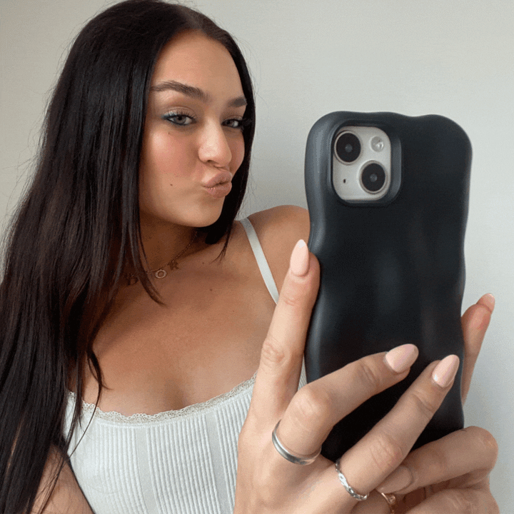 A woman with long dark hair takes a selfie with her smartphone, showcasing the 3D Boujee Phone Case - Matte Black by Coconut Lane. She wears a white tank top, makes a pouty face, and holds the phone in her left hand against a plain white background.